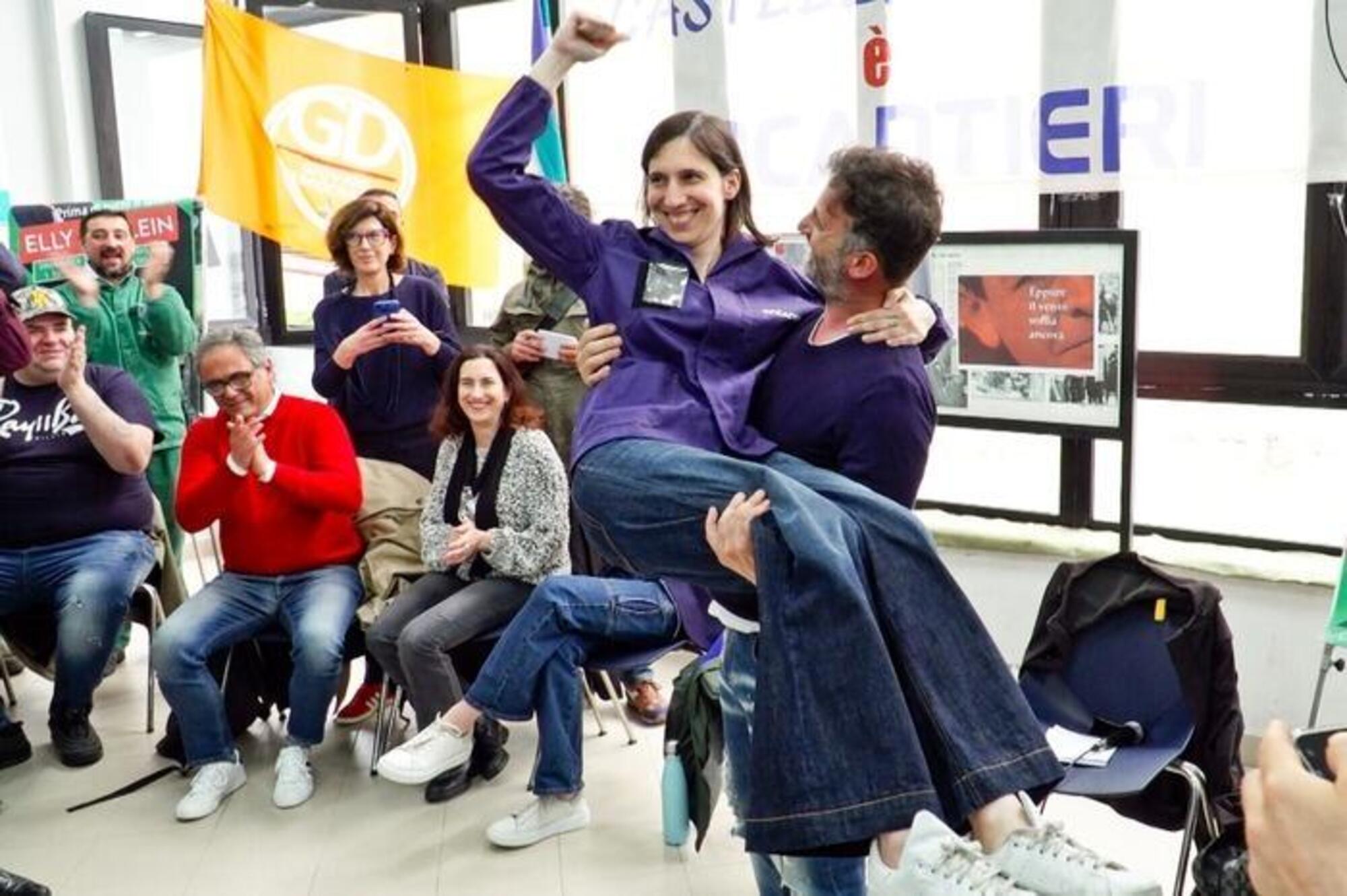 Elly Schlein vestita con una tuta da lavoro presa in braccio da un operaio di Fincantieri